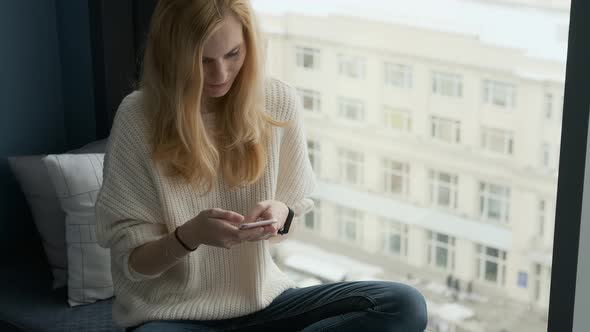 charming blonde woman with smartphone by the window