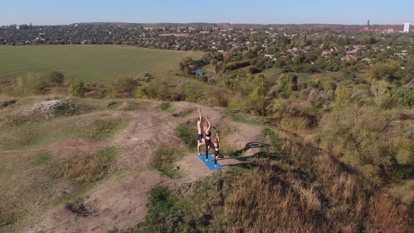 Orbiting Aerial Shot of Family of Three Mother Father and Daughter Do Yoga Exercises on Top of Hill alt