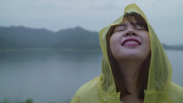 Young Asian woman feeling happy playing rain while wearing raincoat standing near lake. alt