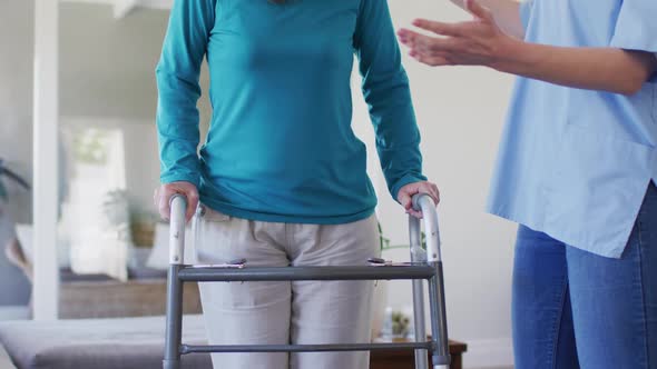 Female health worker assisting senior woman to walk with walking frame at home alt