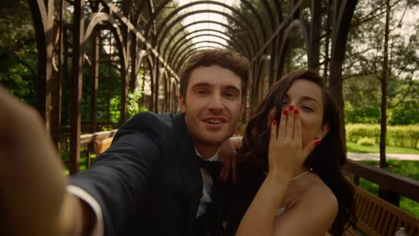 Young Couple Informing About Marriage in Park. Bride Sending Air Kiss on Camera alt