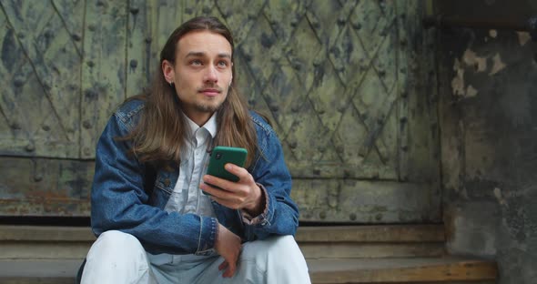 Bearded Millennial Man with Earrings Scrolling News Feed While Sitting at Stairs Outdoors. Young alt