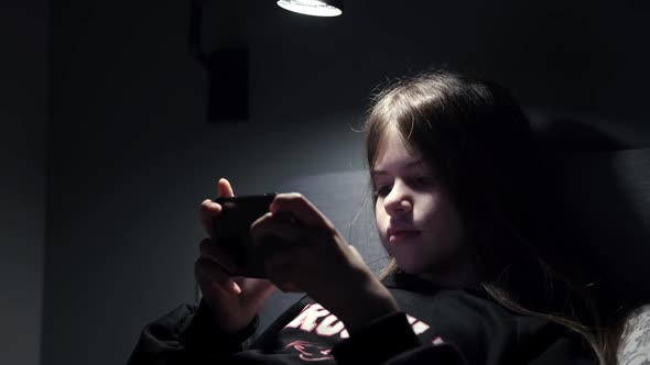 a Little Girl Watches Cartoons Plays Games on a Smartphone at Night in the Dark alt