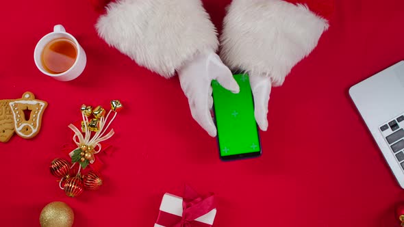 Top View Santa Hands Uses Smartphone and Drinks Tea By Red New Year Decorated Table alt
