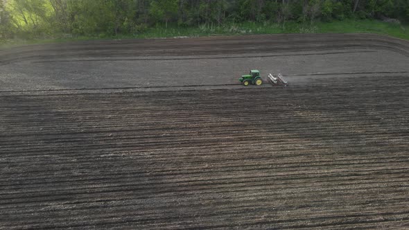 Farmer driving tractor, fertilizing, and seeding a section of farmland. Cloudy sky above the trees.