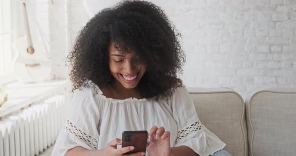 Young Black Smiling Woman Recording Audio Message to Friend Using Smartphone Sitting on Grey Sofa