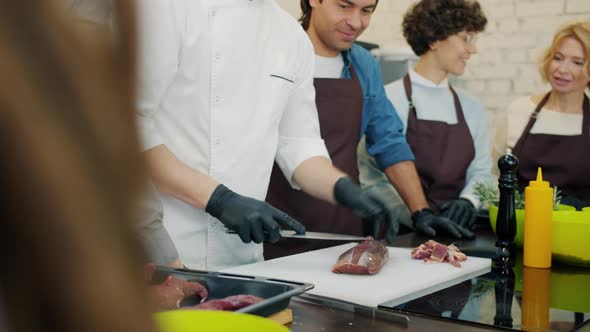 Tilt-up of Professional Cook Preparing Meat Dish in Cooking Class with Cheerful Students alt
