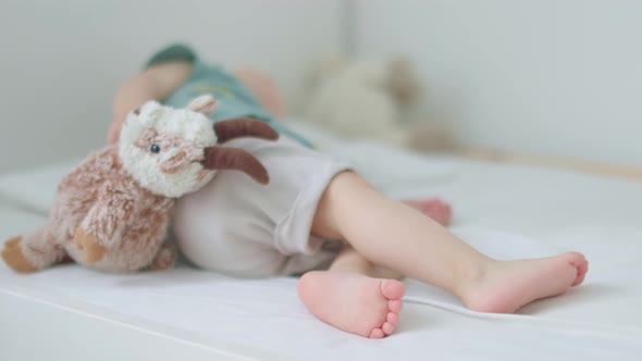 Barefoot Legs of Toddler Baby Lying on Bed with Fluffy Goat in Hands Sleeping alt
