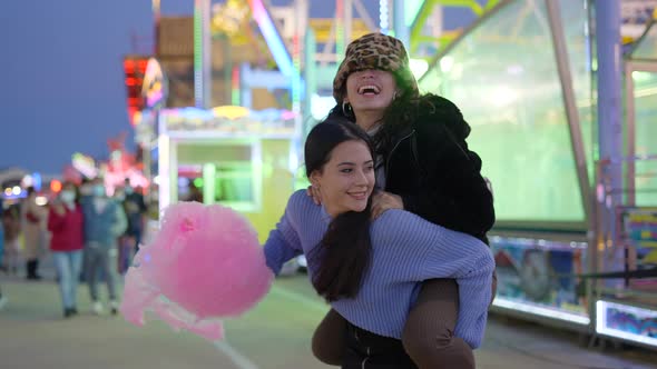 One Girl Gives Another a Piggyback Ride Holding Pink Candy Floss alt