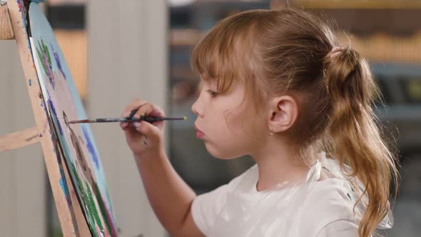 Serious Little Girl Paints with Brush on Canvas on an Easel