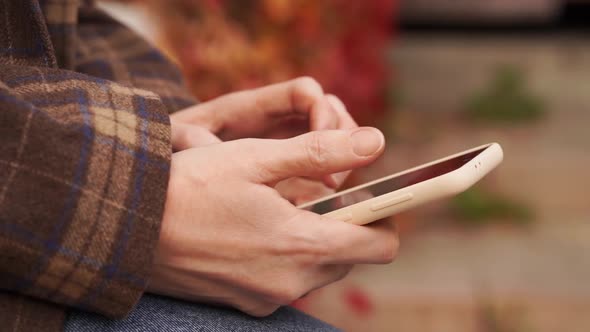 a Woman Holds a Smartphone in Hands and Views Social Media Feed in Autumn Park alt