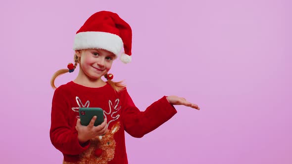 Girl Toddler in Christmas Santa Hat with Mobile Phone Showing Pointing Empty Place Advertising Area alt