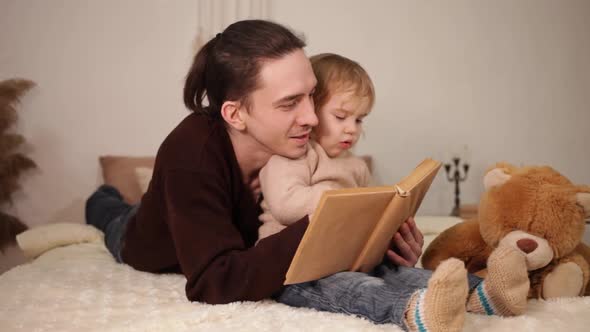 A Dad Reads His Daughter a Book on a Bed in the Room