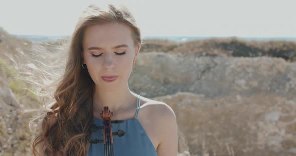 Elegant Woman Holds a Violin Opens Eyes and Looks Into Camera Among Cliffs alt