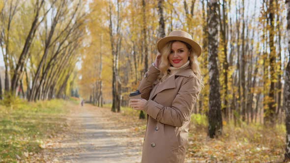 Young Woman in a Brown Coat and Hat Walks in the Autumn Park alt