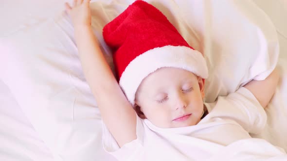Little boy in Santa hat sleeps in a white bed. New year, holidays, christmas. Morning happiness.