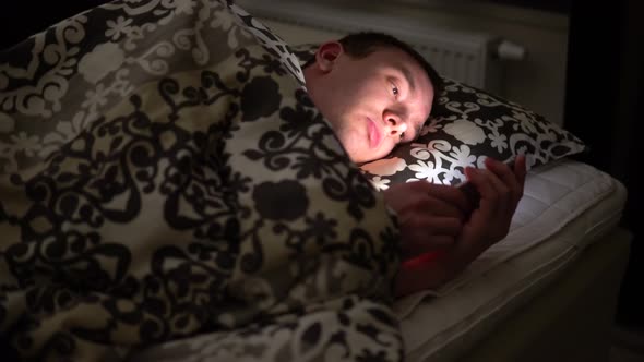 Young Man Uses His Smartphone in Bed Before Sleep. The Light From His Phone Illuminates His Face alt