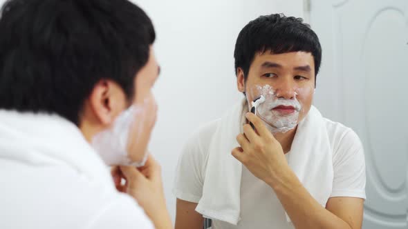 man using razor to shaving his face with cream foam in the bathroom mirror alt