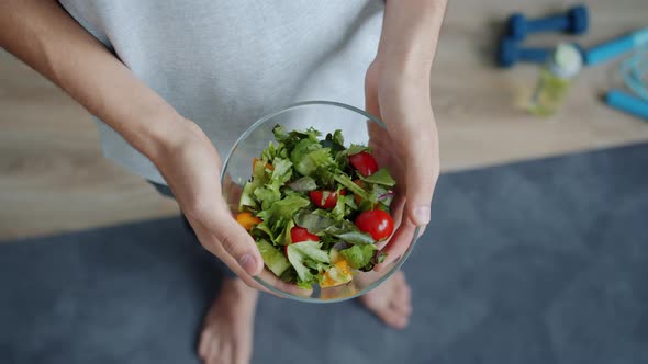 Top View of Man's Hands Holding Bowl of Vegetable Salad Standing on Yoga Mat at Home alt