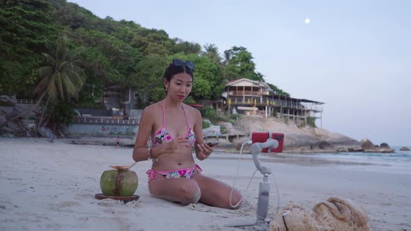 Thai Girl Blogger in Bikini Broadcasting Live Stream Sitting on White Sand Beach at Sunset alt