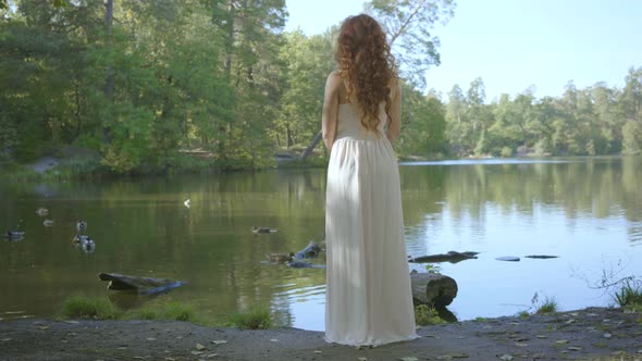 Back View of Beautiful Slim Woman with Red Hair in White Long Dress Standing on the Riverbank alt
