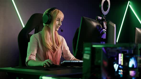 Young Gamer Girl Wearing Pink Clothes and Headset Sitting in Gaming Chair By a Desk Focused on alt
