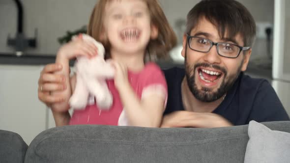 Little Child Girl with Father Fun Plays Jumps From Behind Sofa with Toy Scare Scream and Laugh