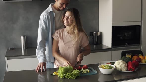 A Young Man is Kissing His Girlfriend When She's Making a Fresh Salad for Them alt