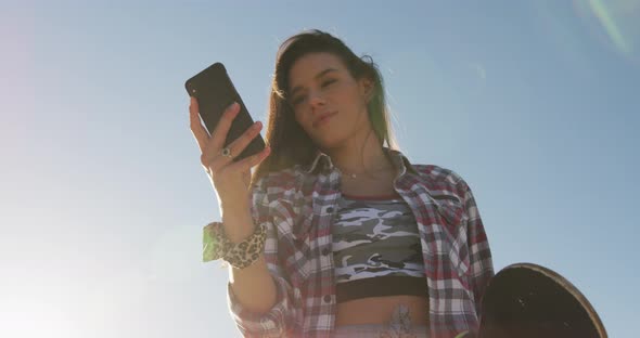 Smiling caucasian woman using smartphone and holding skateboard at a skatepark alt