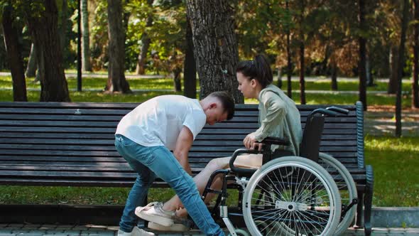 A Couple Walking in Park  a Guy Transfers Woman From a Wheelchair to Bench alt