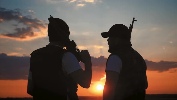 Silhouettes of Two Soldiers with Weapon Against a Sunset alt