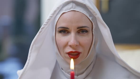 Closeup Face of Gorgeous Woman in Nun Costume Blowing Out Candle Looking at Camera Winking and alt