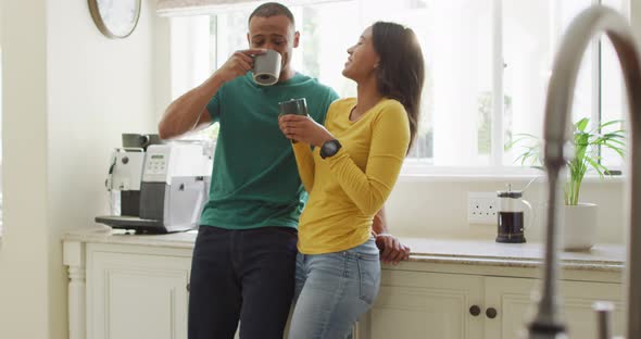 Happy biracial couple standing in kitchen, drinking coffee and laughing alt