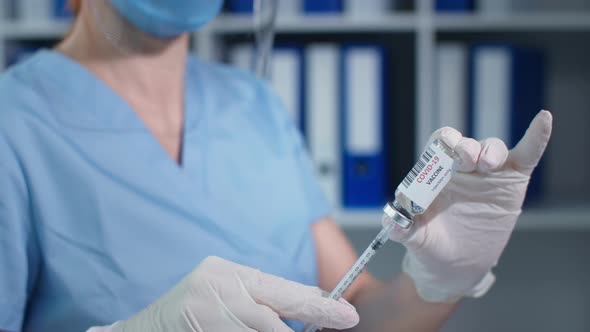Masked Doctor Closeup in Blue Uniform and Protective Gloves with Syringe Pierces the Rubber Seal of