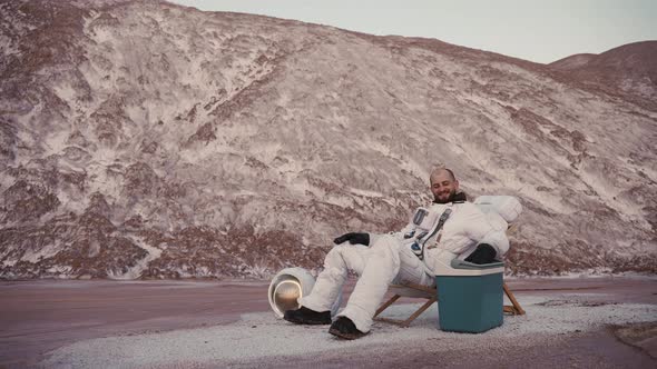 Astronaut Resting in a Chair By the Hills Without Helmet Drinking Cold Beer alt
