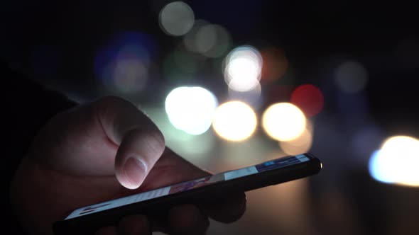 Hand of a Man With a Smartphone in the City at Night Against the Background of Blurry Lights of Cars alt