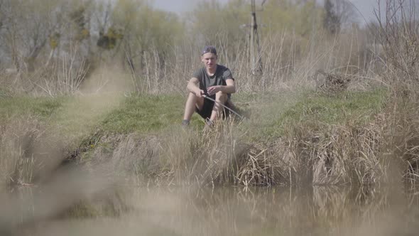Wide Shot Portrait of Focused Young Caucasian Fisherman Sitting on Bushy Lake Shore and Looking at alt