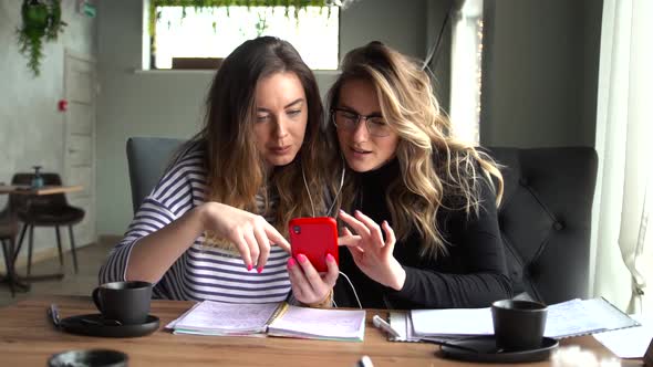 Two Women Sit in a Cafe During the Day and Watch a Video Together on a Smartphone.