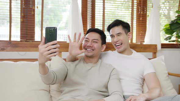 Happy young asian gay couple sit couch use smartphone facetime video call with friends and family