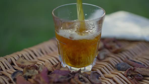 Closeup Apple Juice Pouring in Glass Outdoors with Dried Fruits Lying on Picnic Basket alt