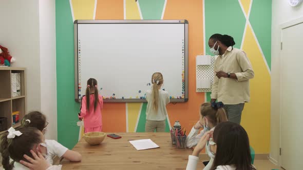 African American Teacher in a Mask Teaches a Group of Masked Children Learning English