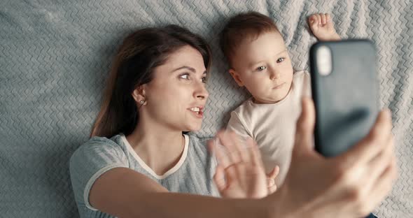 Smiling Woman and Baby Boy Making a Selfie or Video Call in a Bed and Waving Their Hands alt