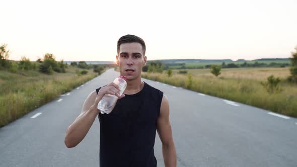 Young Sportsman Walking to Camera Looking Into It Drinking a Bottle of Water alt
