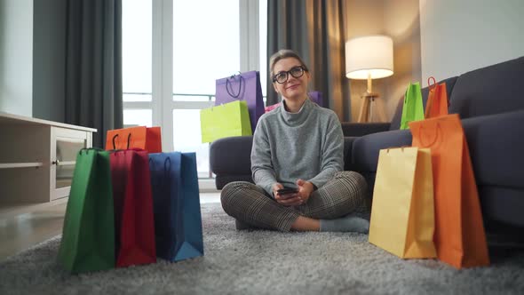 Portrait of a Happy Woman Sitting on a Carpet in a Cozy Room Among Shopping Bags alt
