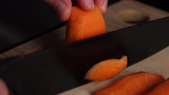 Chopping thin slices from a carrot with a black knife on a wooden cutting board. alt