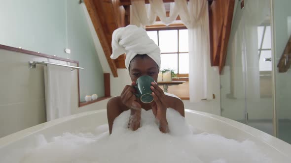 African american attractive woman relaxing in foam bath and drinking coffee in bathroom alt