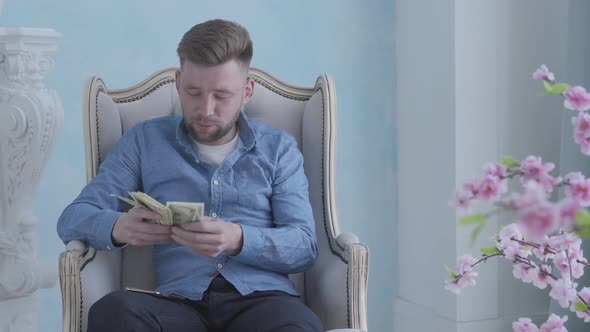 Happy Rich Bearded Man in Blue Shirt Sitting in the White Armchair in the Light Room Counting Money alt