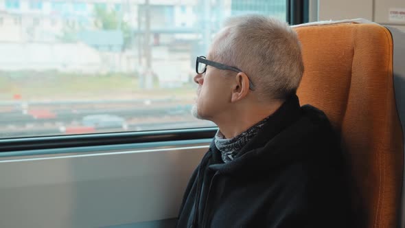 An Elderly Man Travels Sitting on a Train and Looks Out of the Window at the Road with Interest