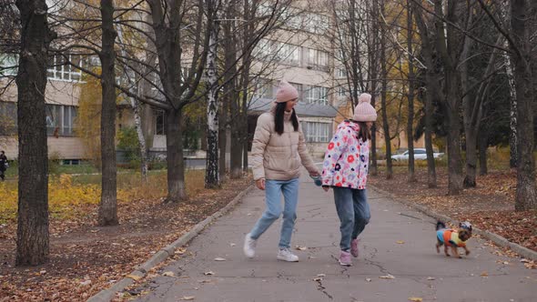 Mom and Daughter are Walking Along an Autumn Alley with a Dog alt