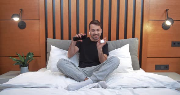 Happy Man Sitting on the Bed in the Bedroom and Listening To Music in Wireless Headphones Positive alt